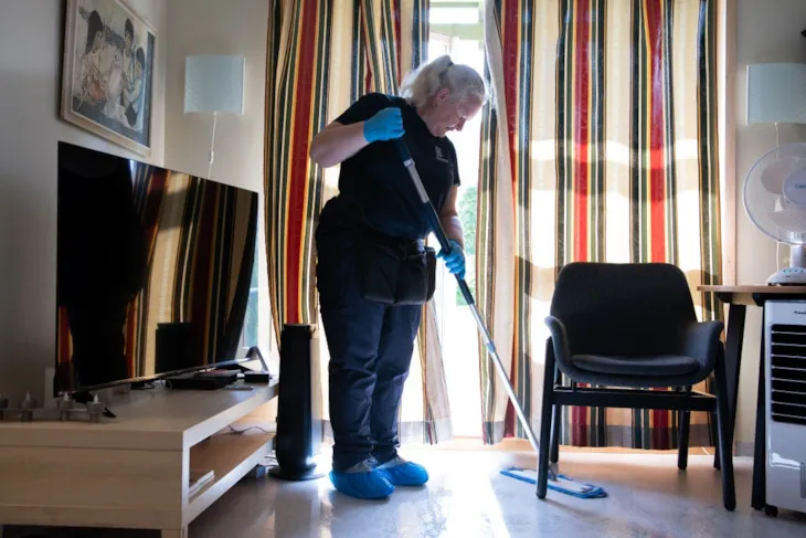 Renholder Aina Remander vasker gulv med mopp. Hun er inne i en leilighet og st&aring;r foran verandad&oslash;ren. Sola lyser inn mellom gardinene. Bak henne er de en tv, og foran henne en stol.