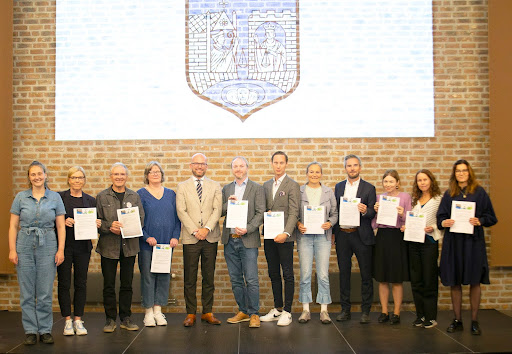 En gruppe på elleve personer står på et podium og holder klimakontrakten. Bak dem er det en vegg med rød teglstein og en prosjektert lys skjerm som viser et Trondheim kommune sin logo.