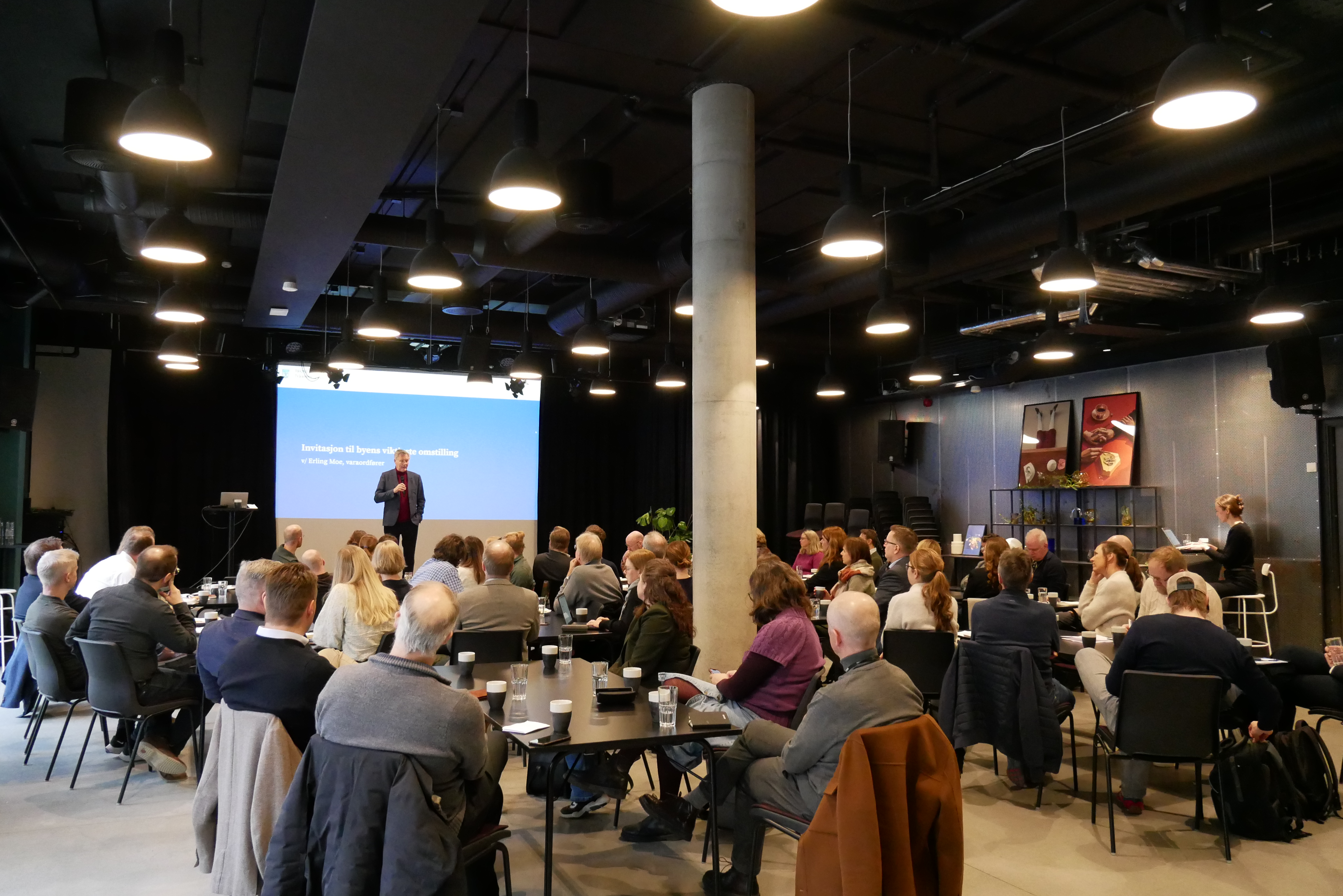 En mann holder en presentasjon for et sittende publikum i et rom med mørkt tak og lyse lamper. En stor skjerm viser tekst bak presentatøren. Publikum sitter ved svarte bord med kaffe og papirer. En søyle står i rommet, og det er dekorasjoner på bakveggen.