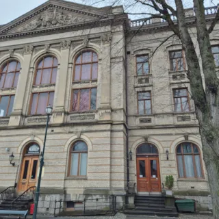 Et bilde av Frimurerlogen som viser en stor, historisk bygning med lys murstein, utsmykkede detaljer, mange vinduer, to store tredører og en stor, bladløs tre i forgrunnen til høyre.