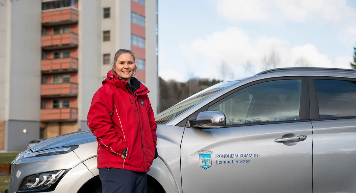 Ansatt i hjemmetjenestens uniform fra Byåsen hjemmetjeneste som står foran tjenestebilen, høyblokk fra Romolslia i bakgrunnen