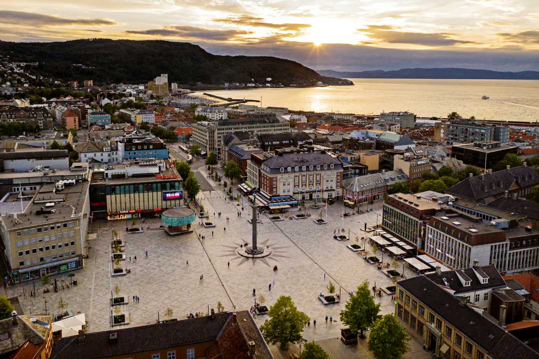Vestvendt dronebilde over Trondheim sentrum med torvet i front og sola over fjorden i bakgrunnen