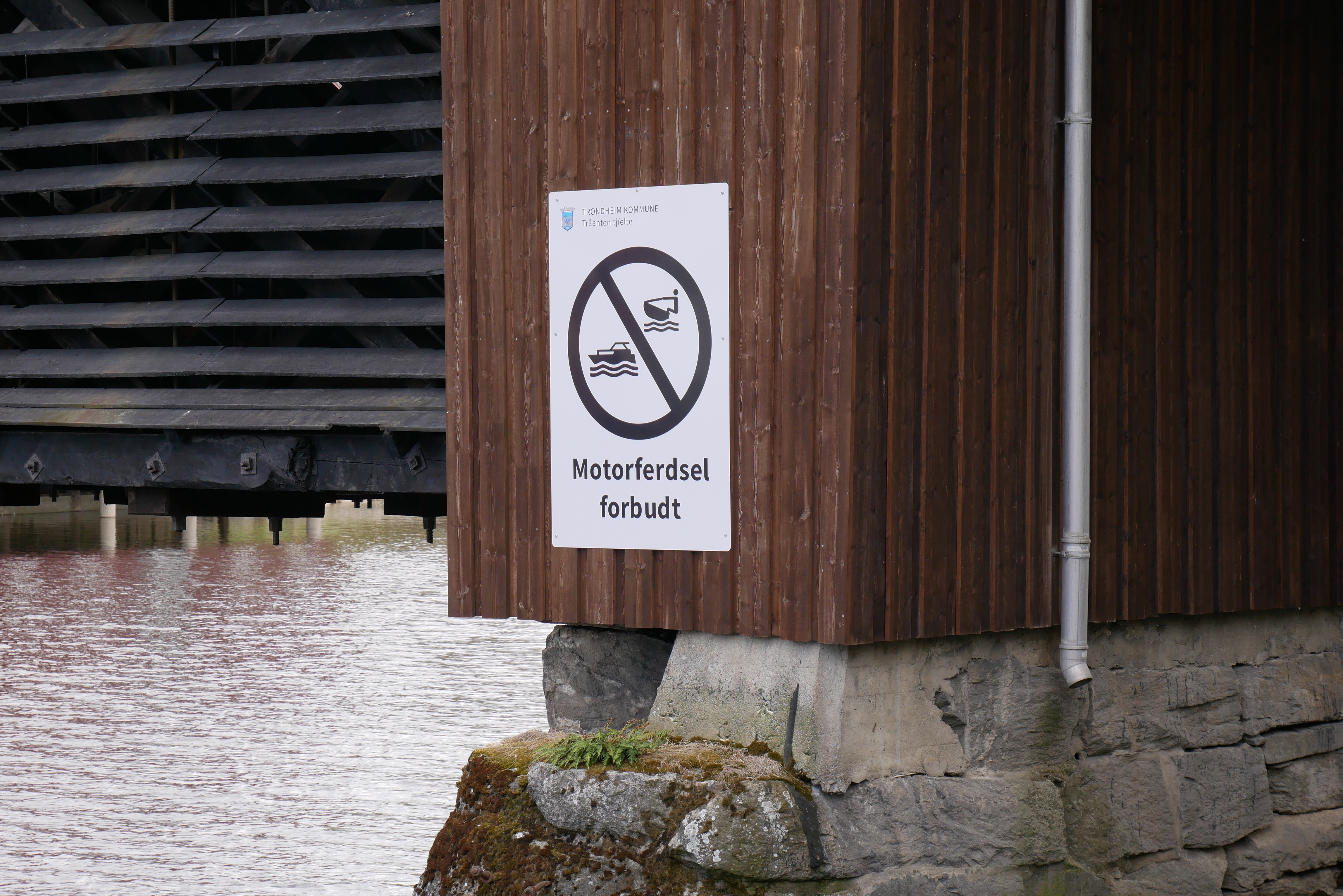 Et hvitt skilt med et forbudssymbol som viser en motorbåt og teksten «Motorferdsel forbudt» er festet til en brun trekonstruksjon, sannsynligvis en bro, over en elv.