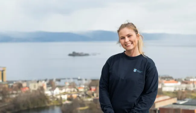 Ung kvinne i blå genser med Trondheim kommunes logo smiler foran uskarp bakgrunn med utsikt mot Trondheimsfjorden og Munkholmen.