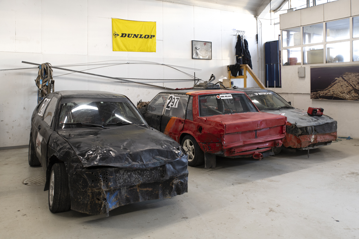 En svart og to røde bilcross-biler inne på verkstedet, med mange bulker.