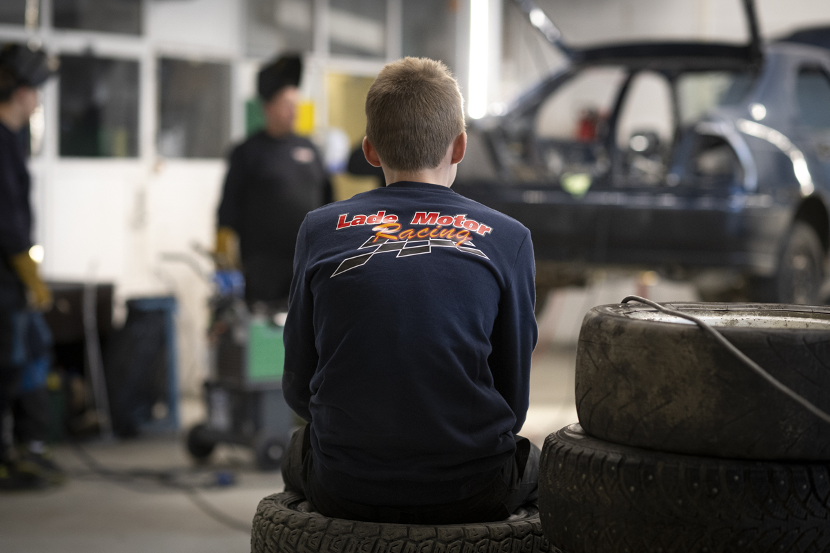 En tenåringsgutt sitter på bildekk med ryggen mot kamera. På genseren hans står "Lade motor" med røde bokstaver.