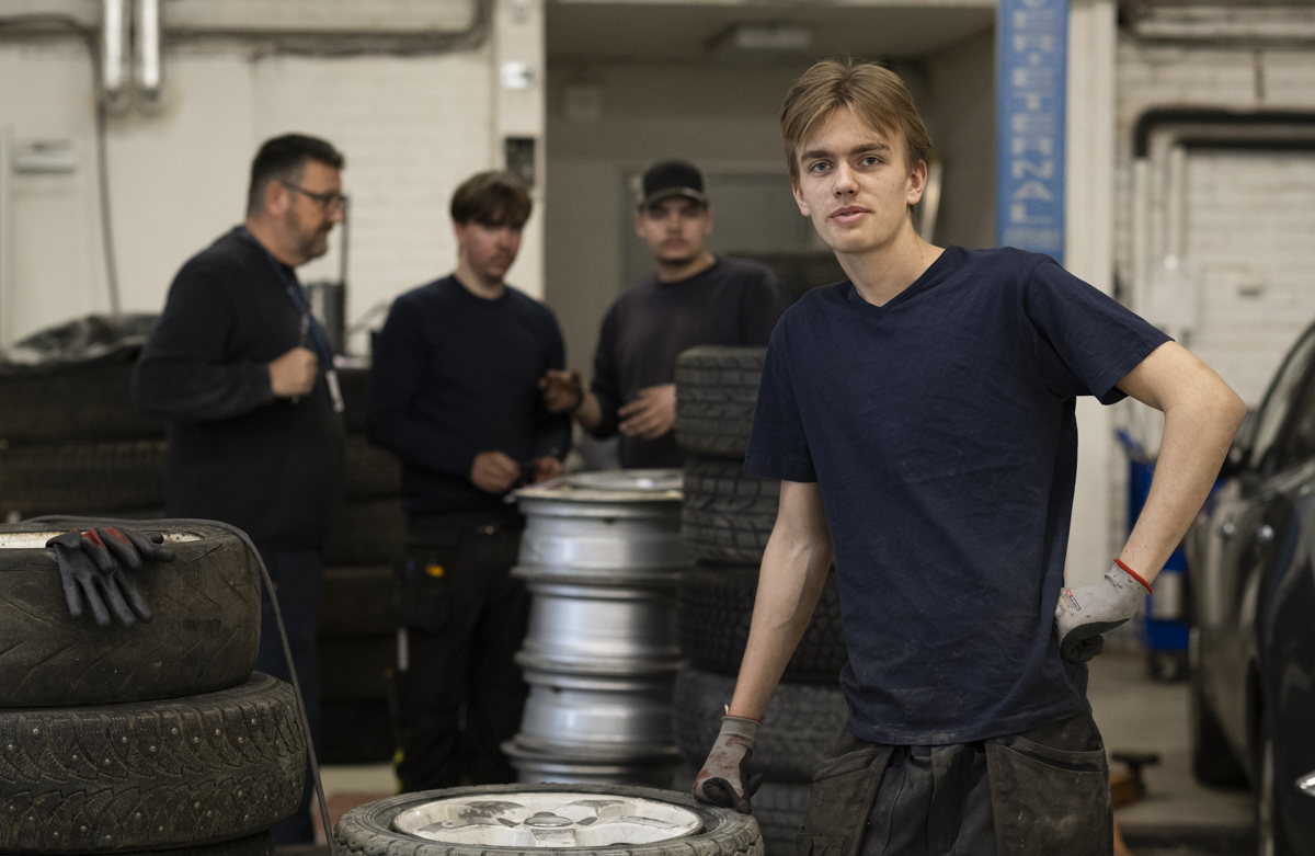 En gutt i slutten av tenårene smiler mot kamera. I bakgrunnen er det stabler med bildekk.