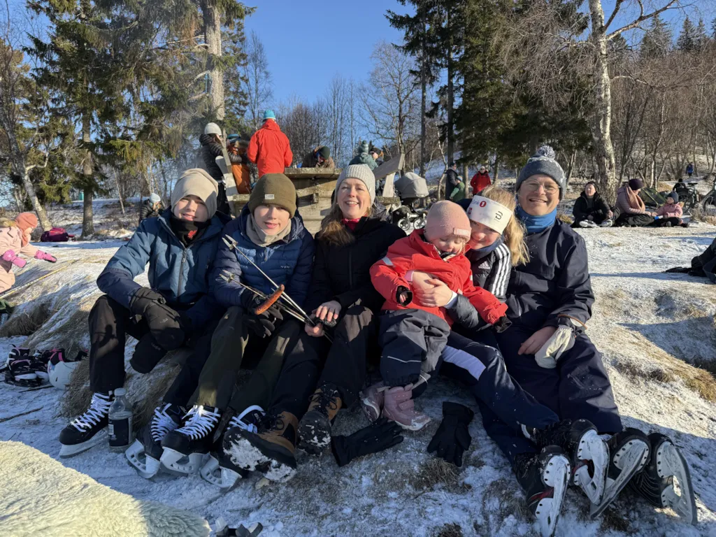 En familie med mor, far og fire barn sitter ved bålet. Alle har på seg skøyter.