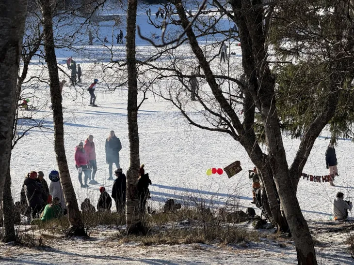 En gruppe folk er samlet ved bål, det henger ballonger i trærne. I bakgrunnen ser man andre folk gå på skøyter.