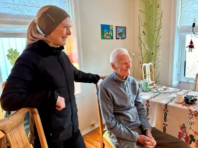 Pasient sitter på en stol med fysioterapeut som står ved siden av stolen.