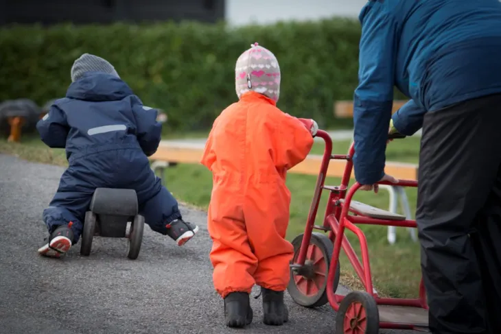 Bildet viser to småbarn og en voksen som leker med sykkel i barnehagen.