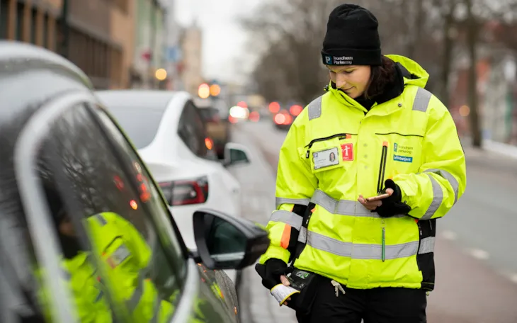 Foto av trfikkbetjent Marie mens hun skriver ut en bot i Kjøpmannsgata. Bakgrunnen er utydelig.