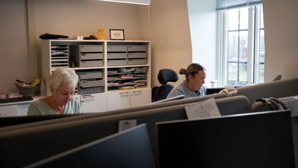 To kvinner sitter i et kontorlandskap og jobber foran pc-skjermer. Vi kan ikke se hva de har oppe på skjermene.
