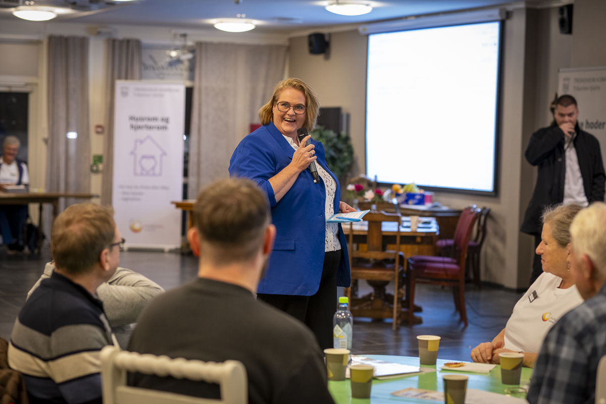 En kvinne i blå blazer snakker i mikrofon og smiler.