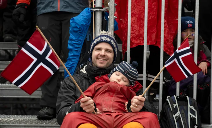 En mann sitter med et sovende barn på fanget. Han smiler mot kamera og har norske flagg i hendene.