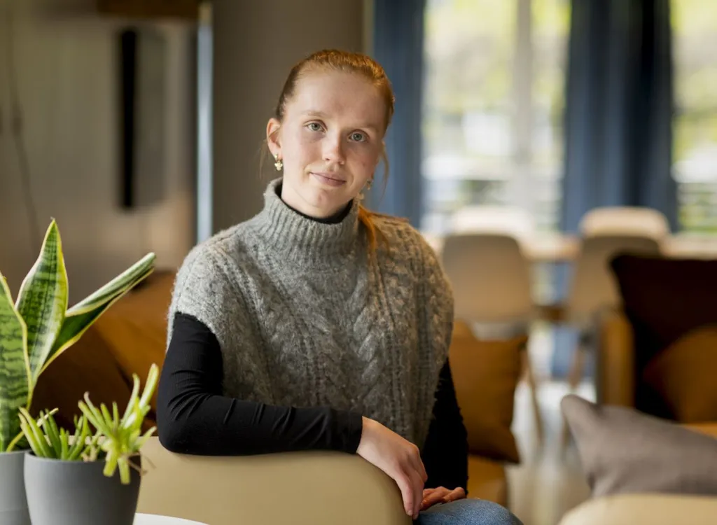 Lærling Thea Udness sitter på en stol i stua på Byåsen BOA. Bakgrunnen er diffus, mens Thea ser i kamera. Vi ser den øvre delen av kroppen hennes. I forkant ser vi planter i venstre hjørne.