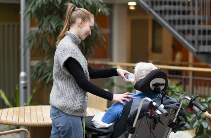 Thea står ved siden av pasienten som sitter i rullestol Hun holder en drikkekopp og hjelper beboeren med å drikke.f
