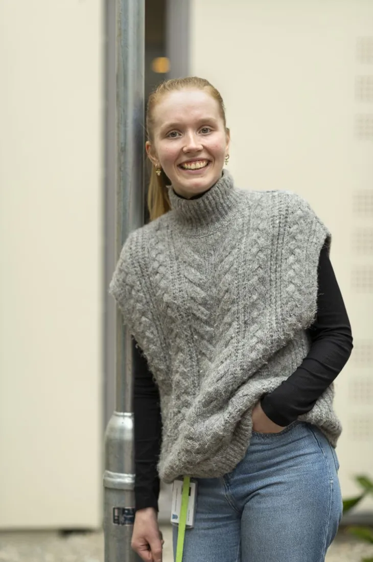 Thea stående lent inntil en lyktestolpe. Hun har venstre hånd i fremre bukselomme.