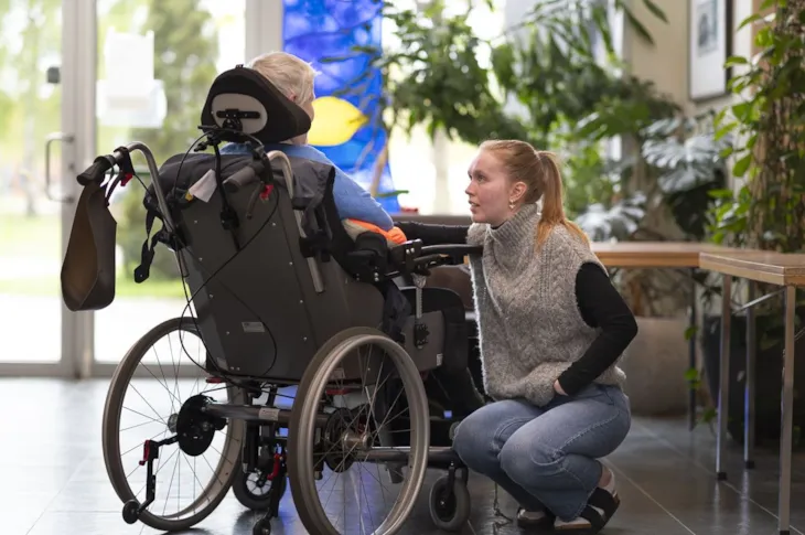 Thea sitter p&aring; huk og snakker med en pasient som sitter i rullestol. Vi ser Thea forfra, mens personen i rullestol sees bakbra.