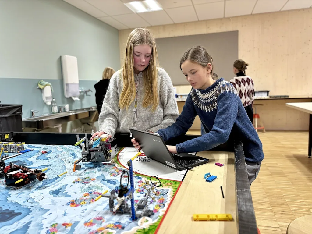 To jenter på 11 år står ved et bord, der bordplata ser ut som en havbunn. Se ser ned på en pc og er konsentrerte i uttrykket.