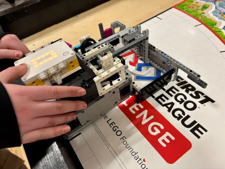 En firkantet robot bygget av lego. To hender holder robotenn og trykker på en knapp som lyser.