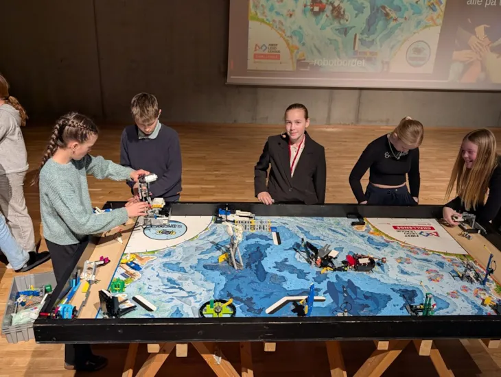 Elevene står ved et bord, der bordflaten ser ut som bildet av en havbunn. Det er flere lego-roboter på bordet som de jobber med.