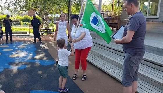 Et barn tar imot Grønt flagg- flagget fra daværende ordfører Rita Ottervik. Flere voksne står rundt dem. Det er sommer og sol.