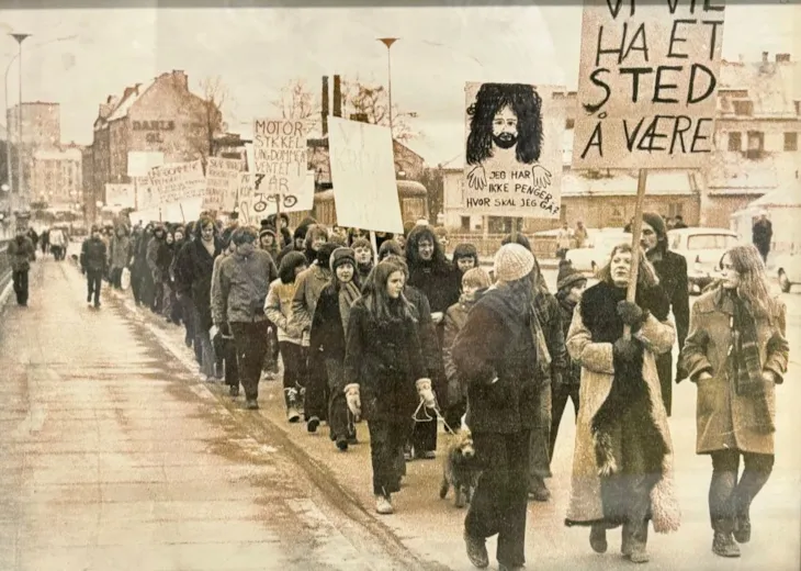 Et gammelt foto av et demonstrasjonstog der de holder opp skilt der det står "jeg har ikke penger hvor skal jeg gå?" og "Vi trenger et sted å være"