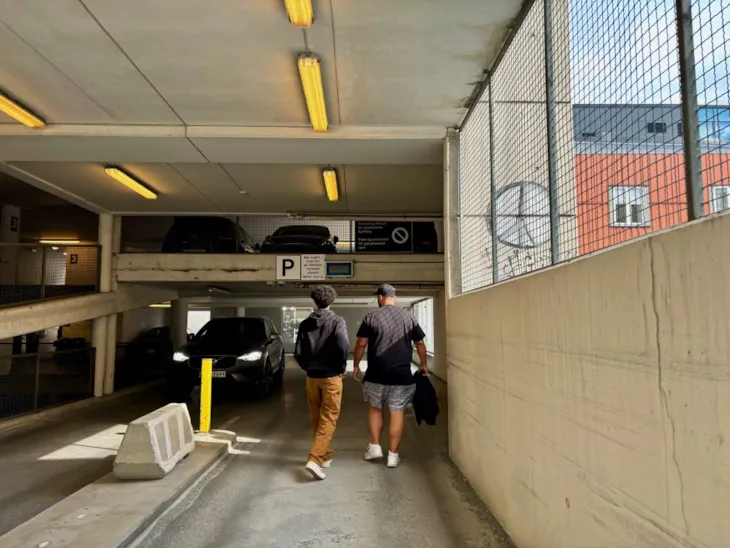 To menn er i ferd med å gå inn i eit mørkt parkeringshus, i det ein bil køyrer ut. Utanfor er det sol og blå himmel.