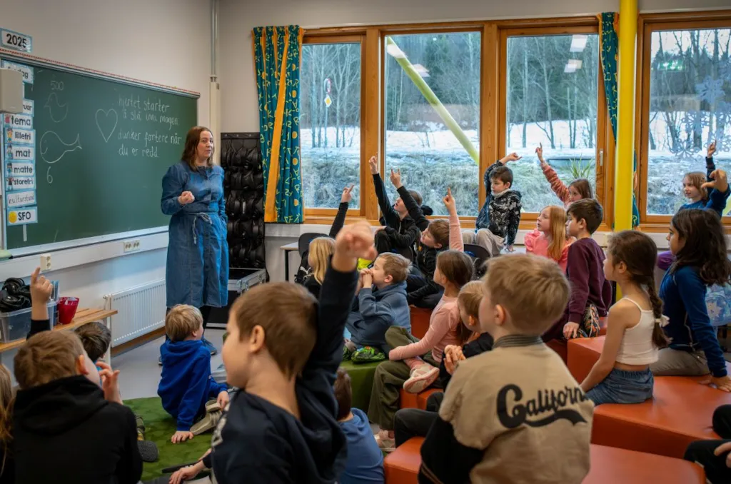 Vi ser ein klasseromssituasjon, der ei gruppe elevar i 8 - 9-årsalederen sitt i gruppe rundt læraren som står ved tavla. Fleirtalet av barna har hånda i veret.
