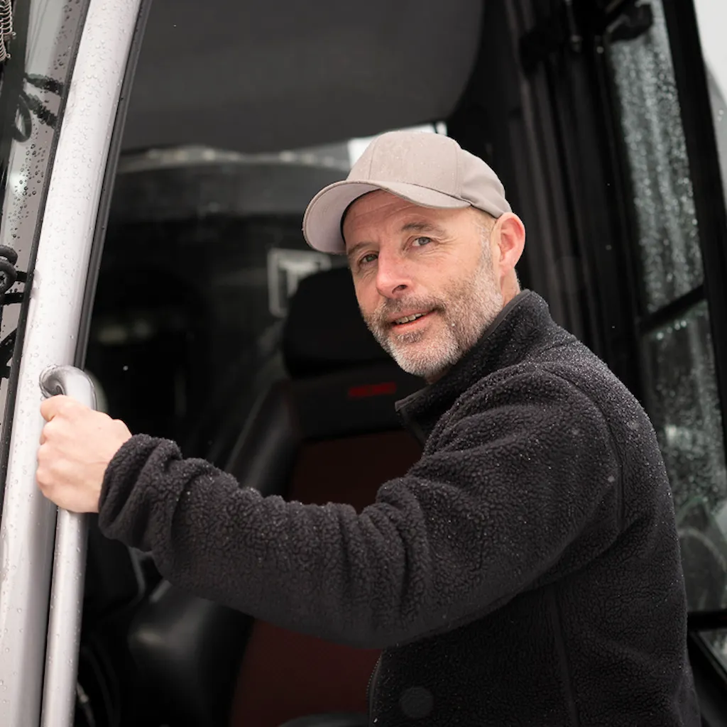 Roger har sorte klær på seg. Står foran løypemaskinen i skogen i Granåsen.