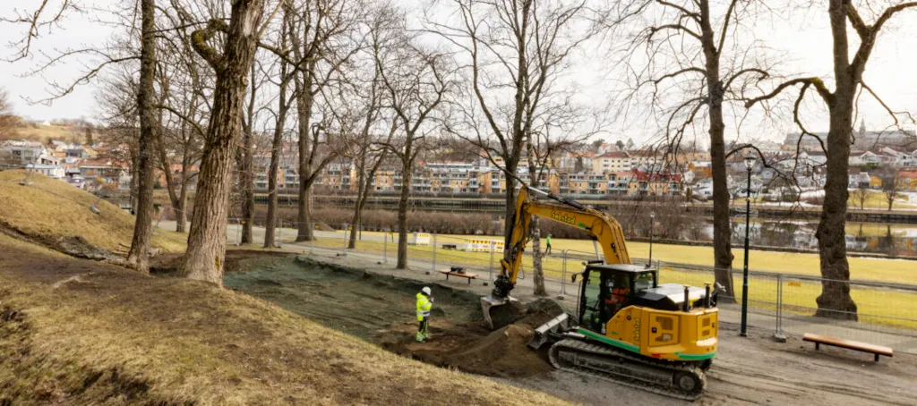 Bilde av en elektrisk drevet gravemaskin som graver på lekeplassområdet på Marinen. Vi ser husrekka på andre siden av Nidelva i bakgrunnen.