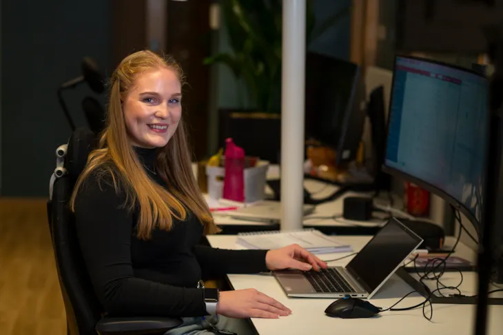 Emilie sitter ved pulten sin og smiler til kamera.