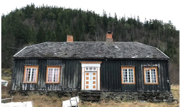 En etasjes bygning med valmtak, mørk trekledning og rikt dekorert portal i hvitt.