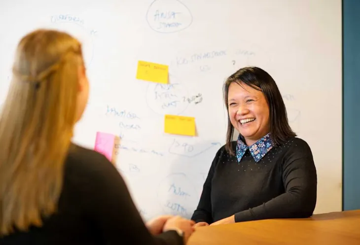 Foto av avdelingsleder Phuong Pedersen sittende ved et bord. I bakgrunnen er det en tavle.