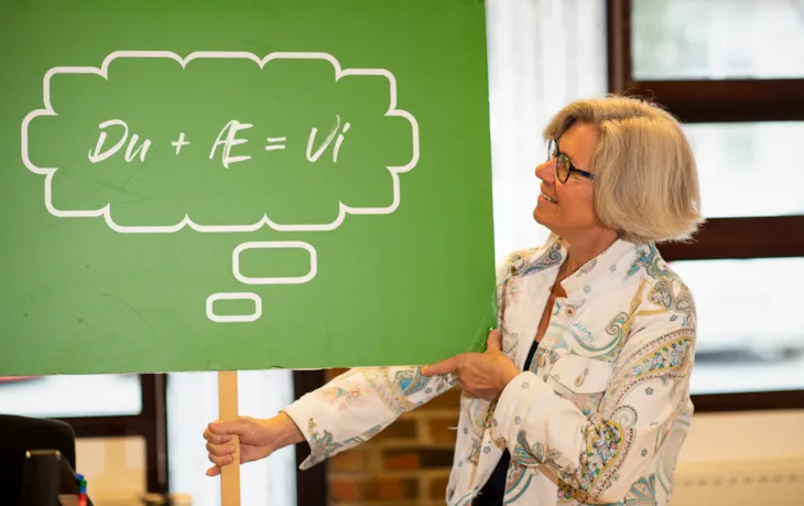Rektor Gro Kristansen holder et skilt med påskriften "Du + æ = vi" inne i et klasserom.