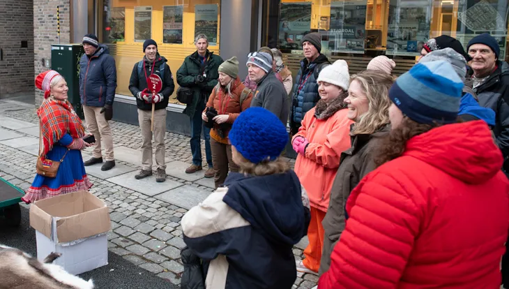 Ei kvinne i kofte snakker til en gruppe på ca 20 mennesker.