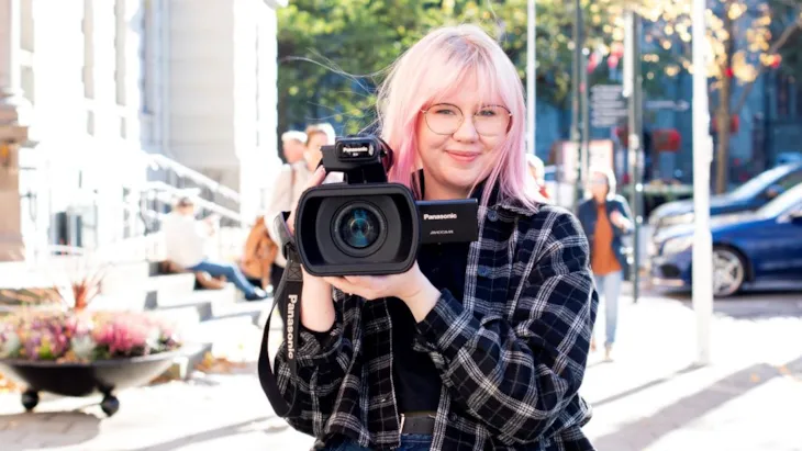 Dame som smiler og holder et videokamera utenfor rådhuset med nidarosdomen delvis i bakgrunnen
