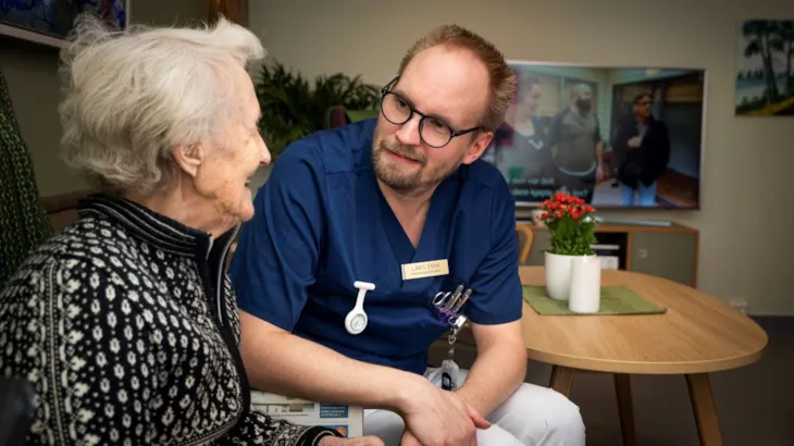 Eldre dame i samtale med mannlig ansatt p&aring; et helse- og velferdssenter. Begge sitter og smiler og ser p&aring; hverandre.