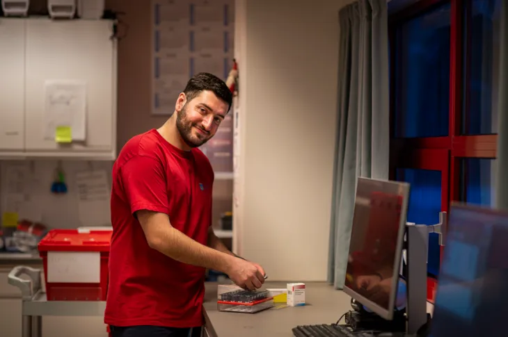 Malik står på medisinrommet og klargjør en medisindosett mens han ser i kamera og smiler.