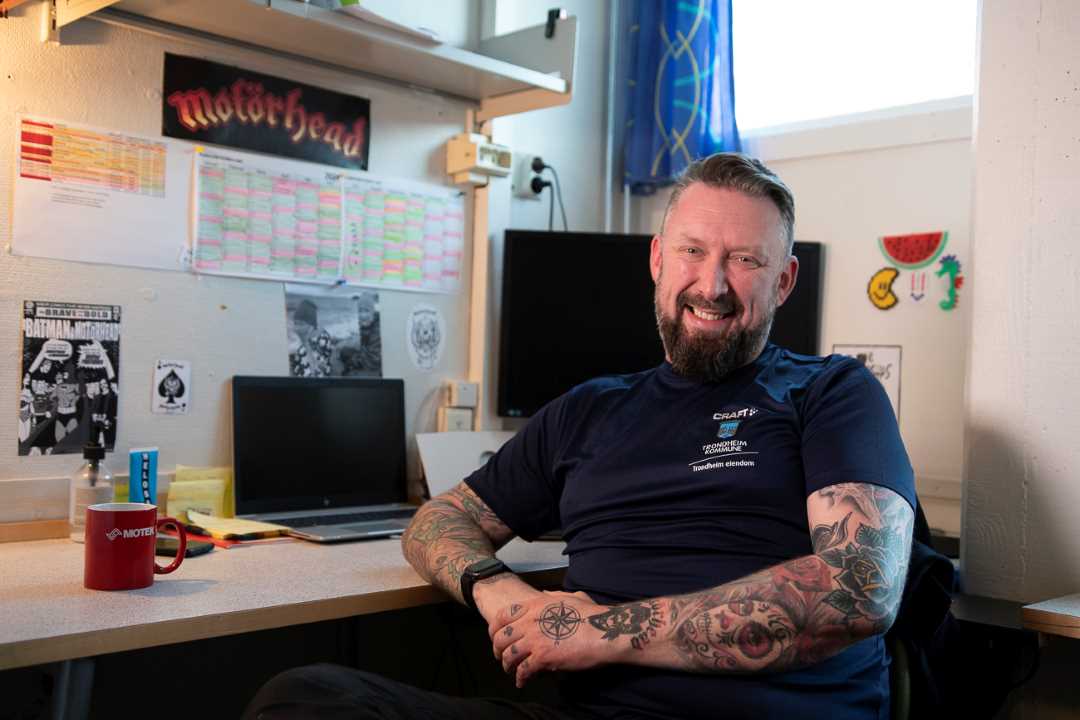 Driftsleder Kim H&oslash;glund Olsen sitter ved pulten og smiler mot kamera. I bakgrunnen er en laptop og en dataskjerm. P&aring; bordet st&aring;r en r&oslash;d kaffekopp.