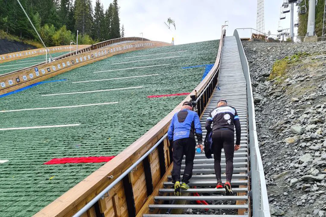 To menn går i trappa ved hoppbakken i Granåsen. En skihopper er i svevet i bakken.