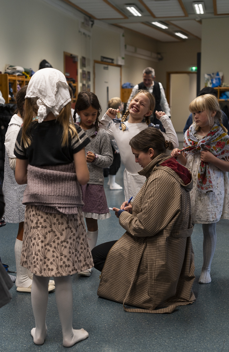En gruppe med jenter i 9-&aring;rsalderen st&aring;r rundt en kvinne som noterer p&aring; blokk. De smiler og er kledd i gammeldagse kl&aelig;r.