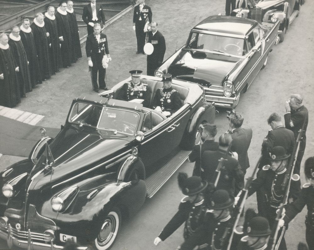 Kong Olav og kronprins Harald på vei til signingen i Nidarosdomen i 1958 i åpen bil.