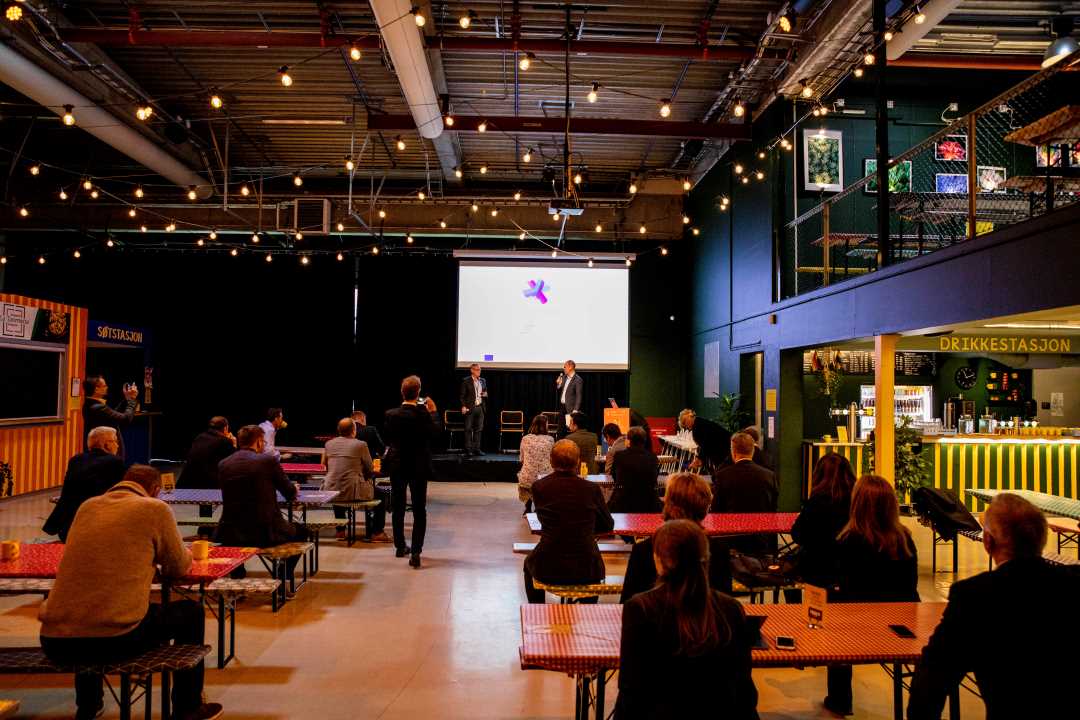 Bilde tatt fra bak tilhørerne på Lager 11 på Sluppen. På den lille scenen står kommunedirektør Morten Wolden og Haakon Skar under partnermøtet for +CityxChange og åpningen av bylab Sluppen.