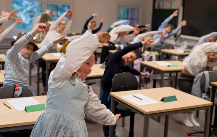 En gruppe elever i småskolen står bak pultene sine, smiler og bøyer seg mot høyre. Barna har på seg gammeldagse klær.
