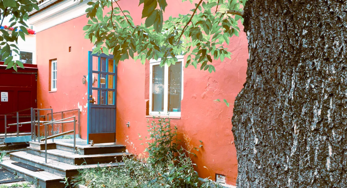 Inngangspartiet til Brovakten. Huset er rødt med blå dør. Vinduene har hvite rammer. Til høyre i bildet ser vi stammen på et tre, som også har grener hengende foran huset.