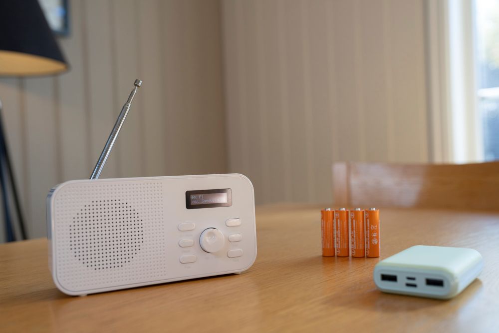 En hvit DAB-radio st&aring;r p&aring; et trebord ved siden av fire oransje batterier og en n&oslash;dlader (powerbank).