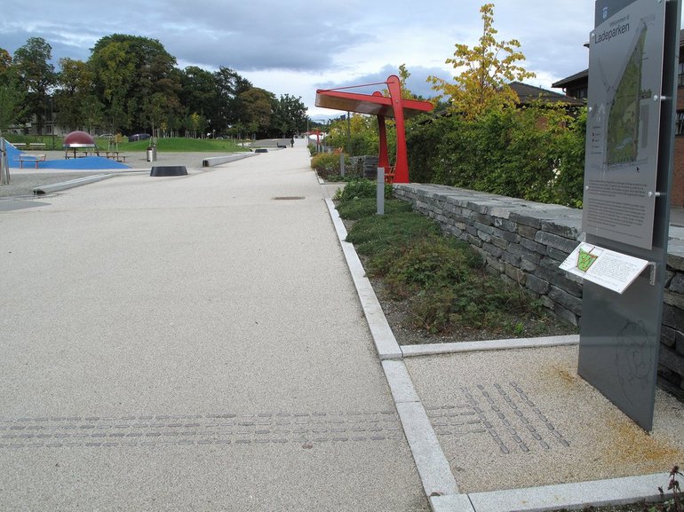 Naturlige og kunstige ledelinjer i Ladeparken gir god orienterbarhet. Taktilt ledelinje og markeringsfelt ved informasjonstavle. Tydelig hvileplass under tak til side for gangsonen