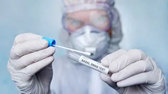 Uklart bilde av person i verneutstyr som holder en nasal swab test i hendene. Hendene og testen er i fokus.